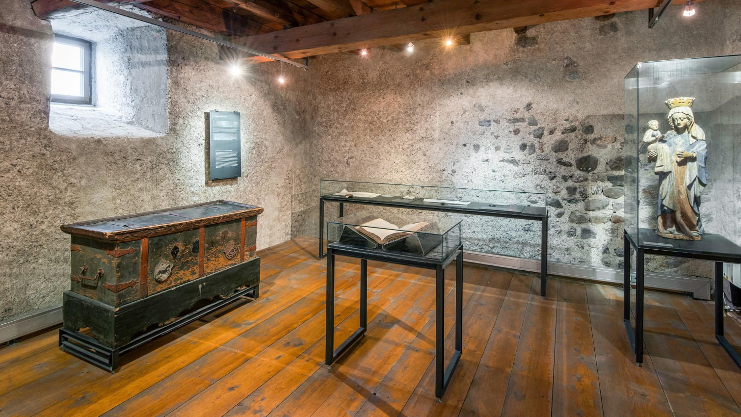 Ein Ausstellungsraum im Museum Kitzbühel mit Holzbalkendecke und antikem Boden. An den Wänden hängen Gemälde, ein Kreuz und eine geö nete Truhe. In der Mitte steht ein Glastisch mit einer alten Karte. 