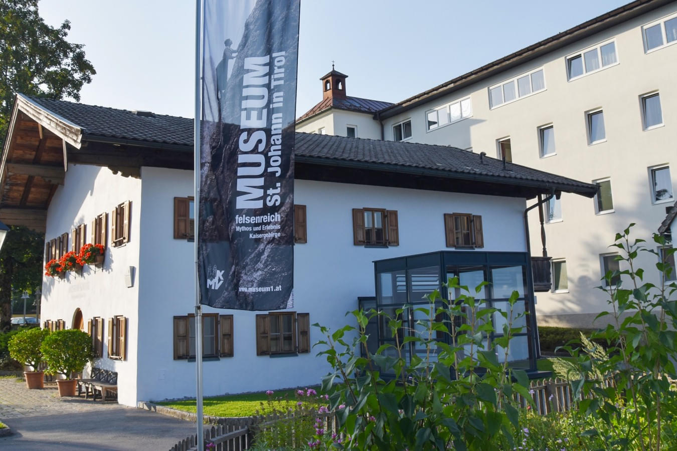 Außenansicht des Museums St. Johann in Tirol, mit einer Fahne, die das Museum bewirbt. Das Gebäude hat eine weiße Fassade mit braunen Fensterläden und Blumenkästen. 