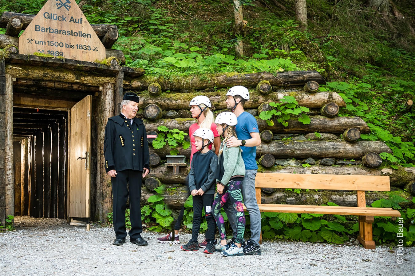 Eingang zum Barbarastollen mit einem Bergwerksführer in traditioneller Kleidung und einer Familie mit Helmen. Im Hintergrund dichter Wald. 