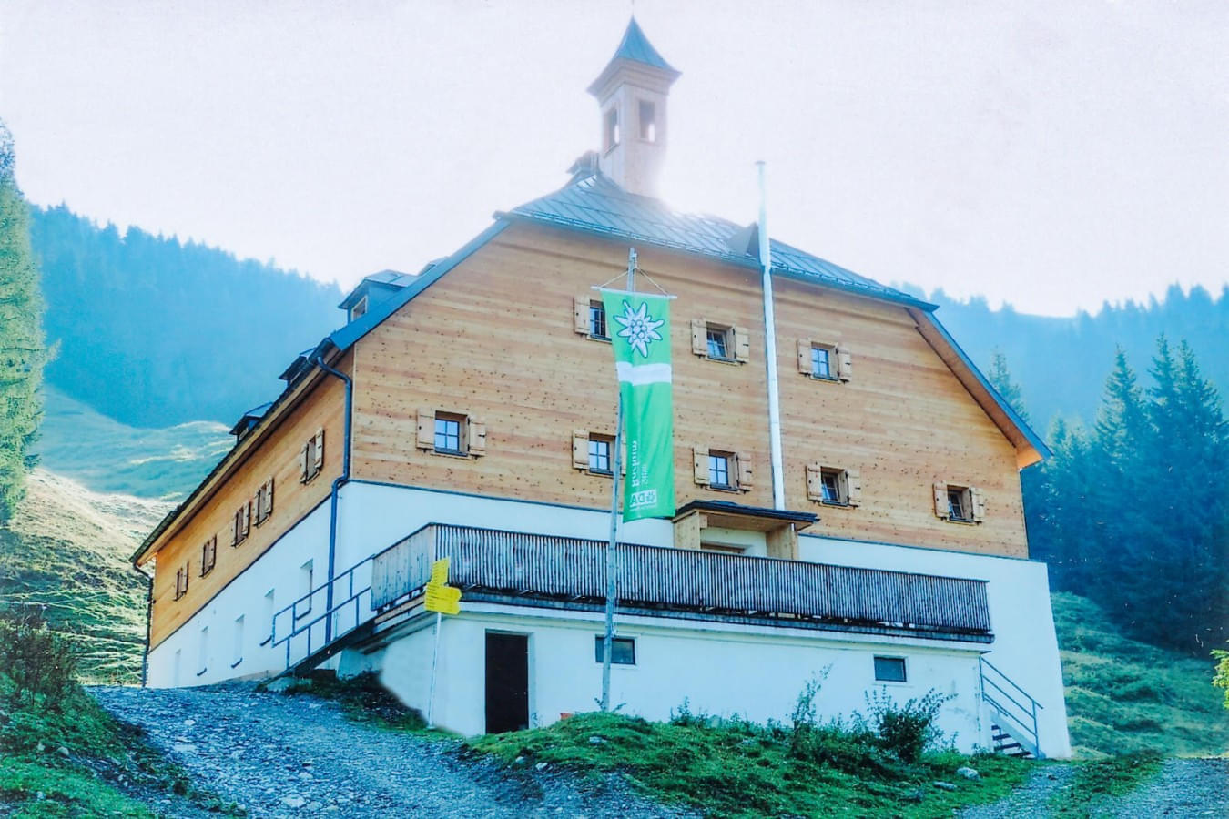 Moderne Aufnahme der Kelchalm mit Schindeldach, davor eine grüne Alpenvereinsfahne und ein Weg, der zum Gebäude führt, umgeben von Bergen und Wald. 