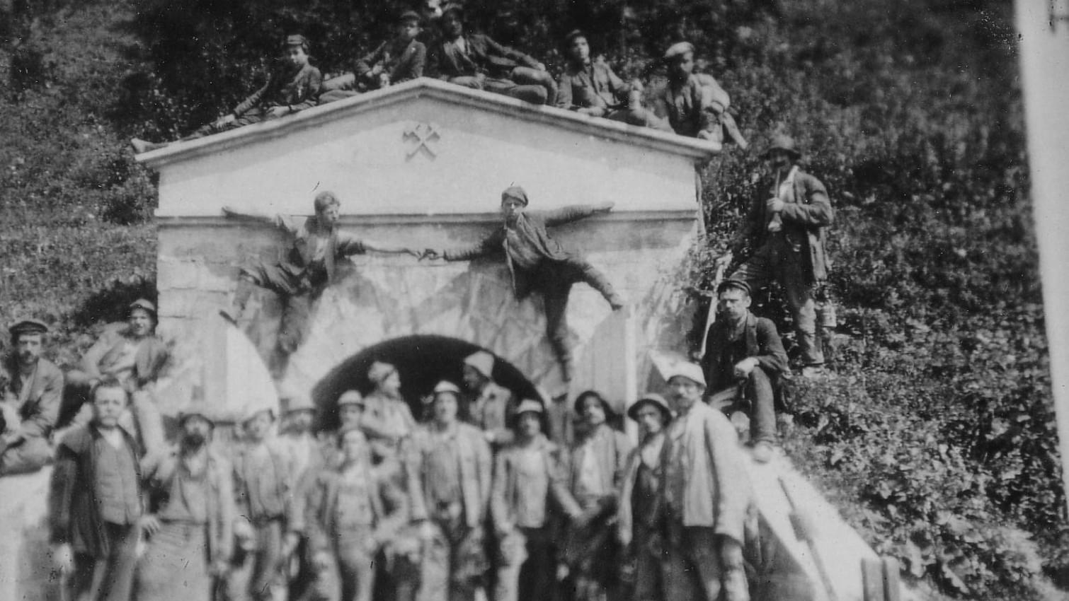 Historisches Foto einer Gruppe von Bergarbeitern vor einem Stolleneingang, einige sitzen auf dem Gebäude. 