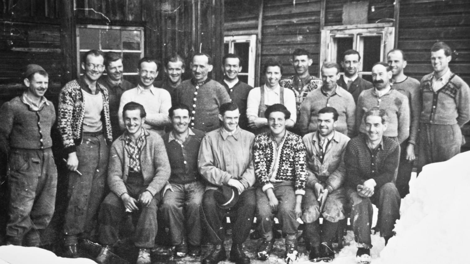 Schwarz-Weiß-Foto einer Gruppe von Bergbauarbeitern in traditioneller Winterkleidung, die vor einem Holzgebäude im Schnee posieren. 