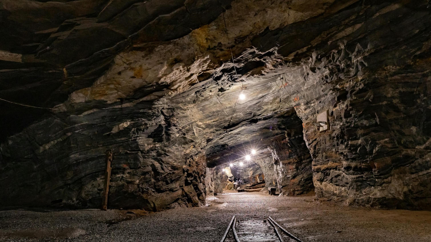 Beleuchteter Bergwerksstollen mit schro en Felswänden, Schienen auf dem Boden - untertage. 