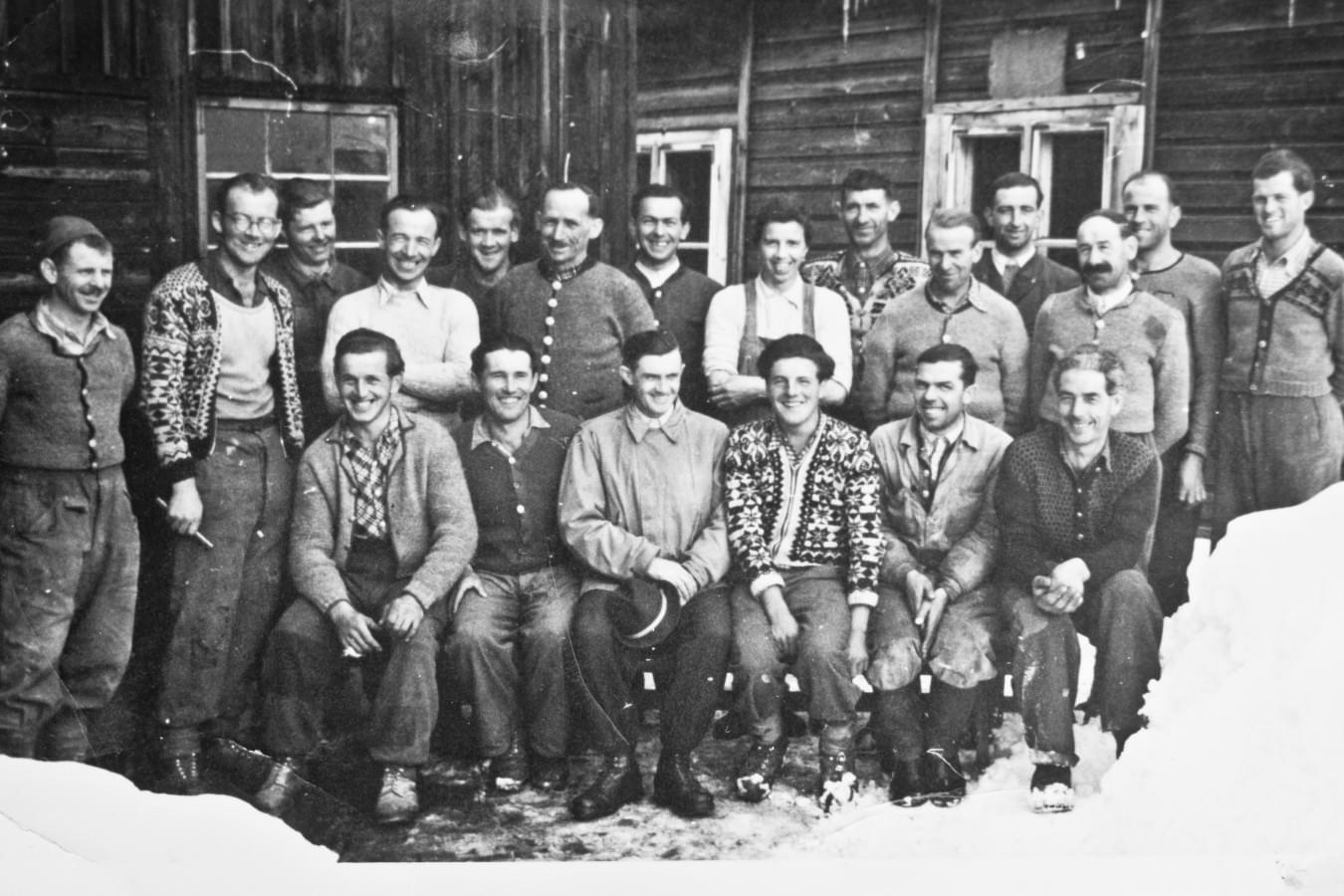 Schwarz-Weiß-Foto einer Gruppe von Bergbauarbeitern in traditioneller Winterkleidung, die vor einem Holzgebäude im Schnee posieren. 