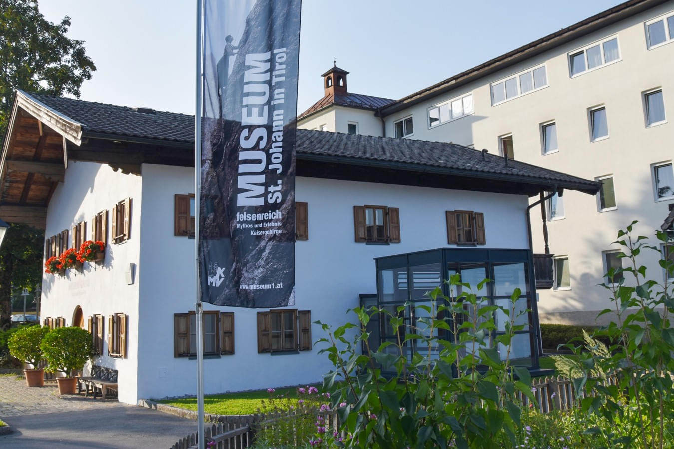 Außenansicht des Museums St. Johann in Tirol, mit einer Fahne, die das Museum bewirbt. Das Gebäude hat eine weiße Fassade mit braunen Fensterläden und Blumenkästen. 