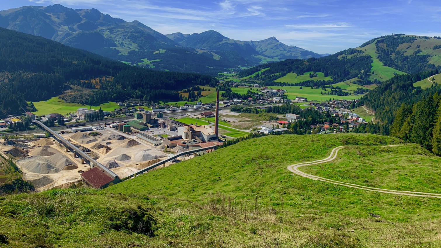 Panoramablick auf das Magnesitwerk Hochfilzen im Tal, umgeben von grünen Wiesen und bewaldeten Bergen unter klarem, blauem Himmel. 