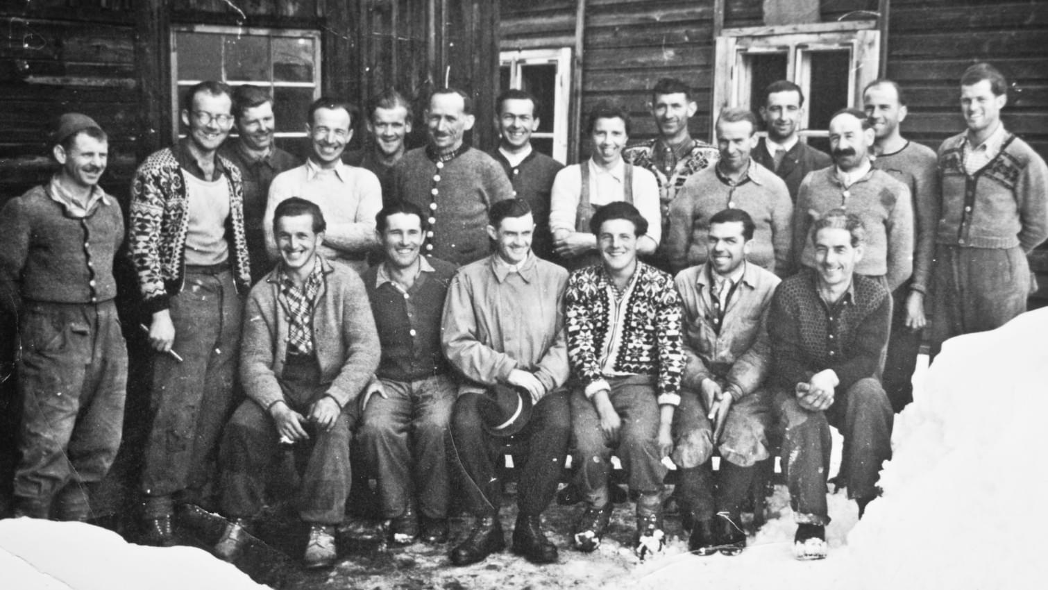 Schwarz-Weiß-Foto einer Gruppe von Bergbauarbeitern in traditioneller Winterkleidung, die vor einem Holzgebäude im Schnee posieren. 