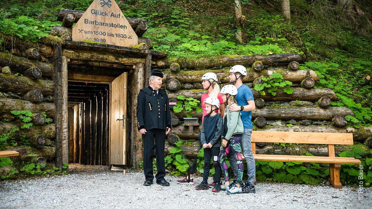 Eingang zum Barbarastollen mit einem Bergwerksführer in traditioneller Kleidung und einer Familie mit Helmen beim Schaubergwerk Leogang.  