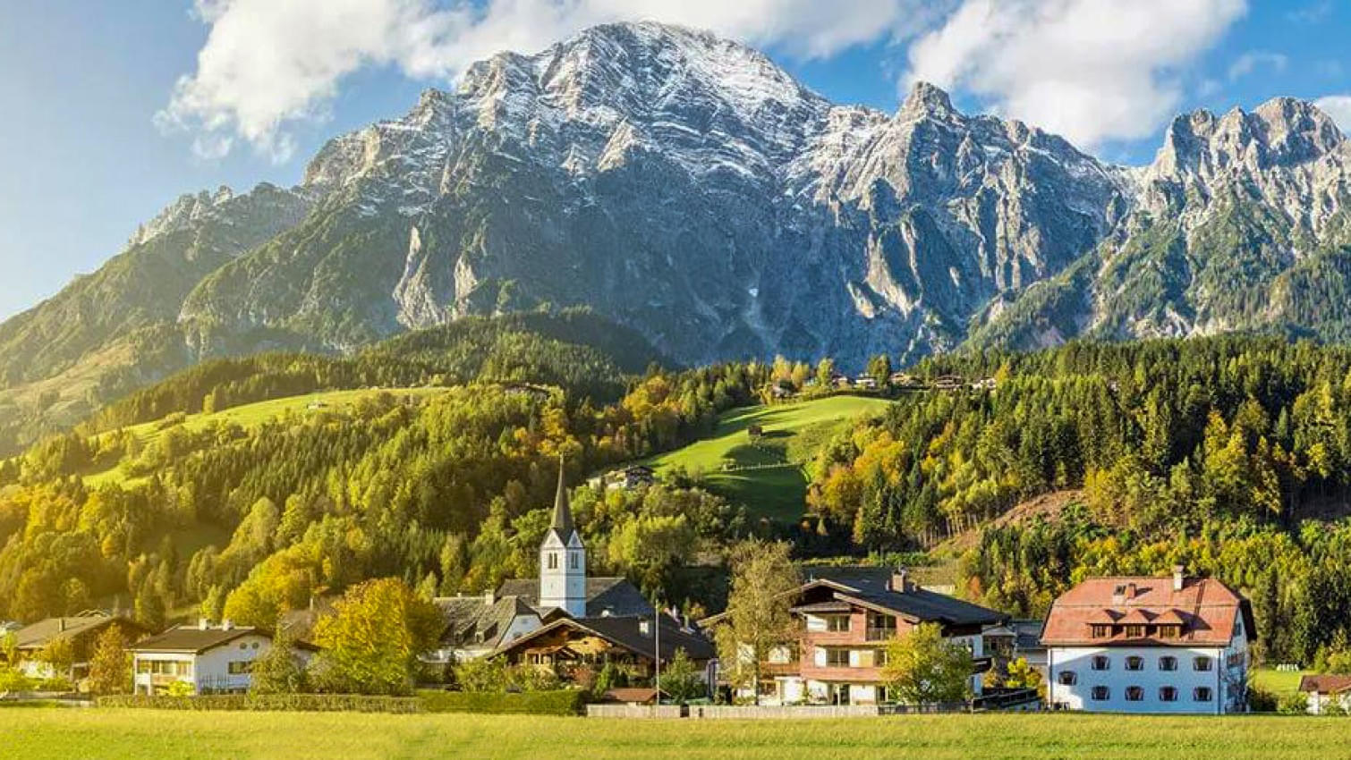 Panoramablick auf die Ortschaft Leogang mit grünen Wiesen, einem Zaun und einem malerischen Tal. Im Hintergrund sind schneebedeckte Berge unter klarem, blauem Himmel zu sehen. 