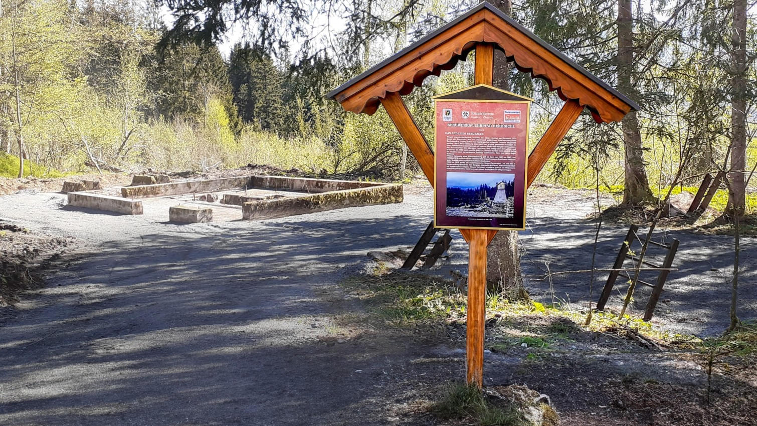 Infotafel des Bergwerks-Lehrpfads Reboichl vor den Grundmauern eines historischen Bergwerksgebäudes in einem Waldgebiet.