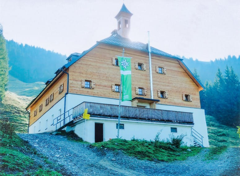 Moderne Aufnahme der Kelchalm mit Schindeldach, davor eine grüne Alpenvereinsfahne und ein Weg, der zum Gebäude führt, umgeben von Bergen und Wald. 