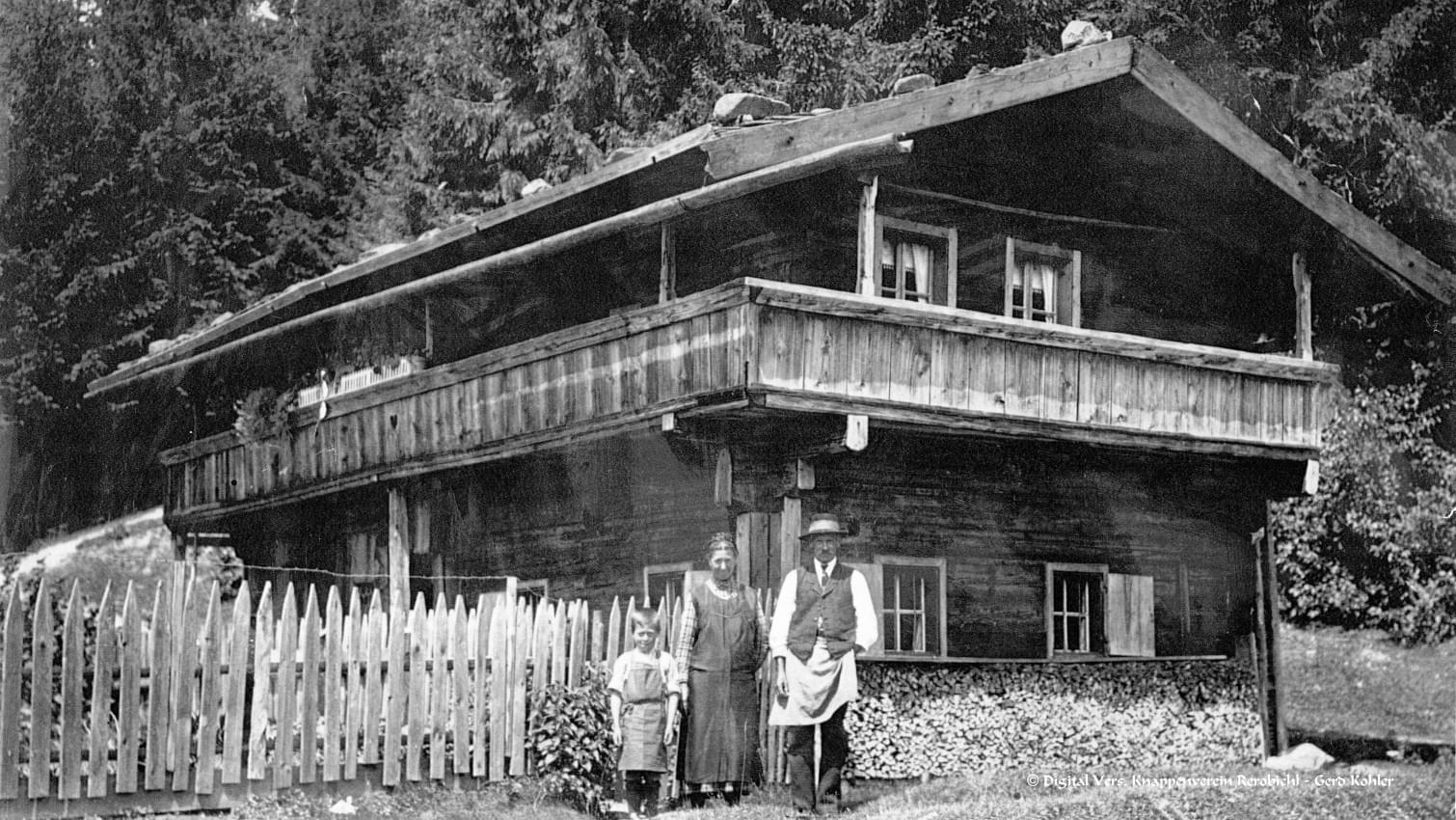 Historisches Foto eines Holzhauses mit Balkon, vor dem ein Mann, eine Frau und ein Kind stehen, umgeben von einem Holzzaun und Wald. 