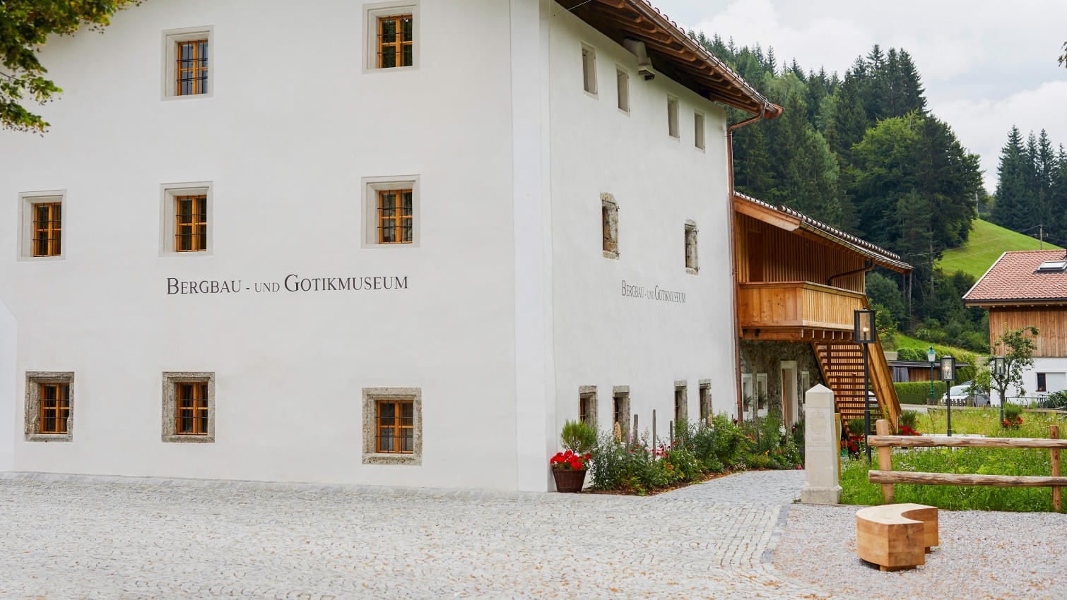 Außenansicht des Bergbau- und Gotikmuseums in Leogang mit weißer Fassade, kleinen Fenstern und einem hölzernen Anbau, umgeben von einer ländlichen Landschaft. 