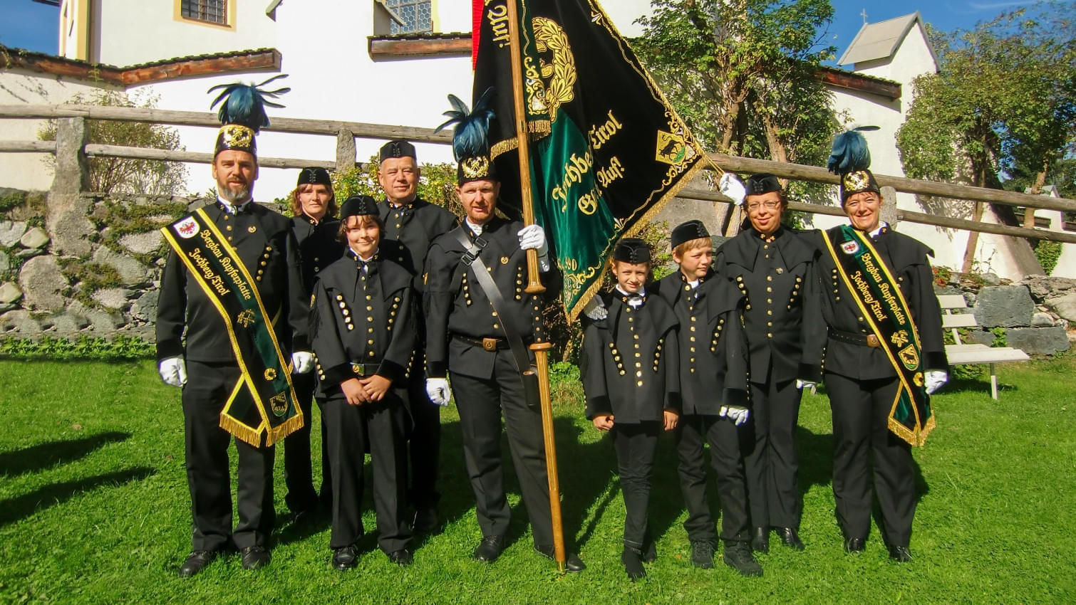 Gruppenfoto einer traditionellen Bergmannsgruppe in festlicher Uniform vor einer Kirche, mit Fahne und Abzeichen.